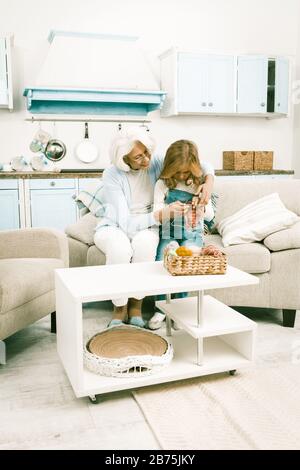 Oma In White Casual Jeans, Die Ihrer Hübschen Kleinen Enkelin Beibringen, Wie Sie Schal Stricken, Während Sie Eng Zusammen Auf Sofa Im Wohnzimmer Mit Ki Sitzen Stockfoto