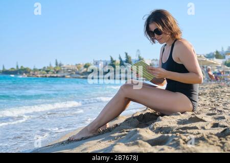 Sommerfrau sitzt auf Sandufer Lesebuch, sonnt sich in untergehenden Sonne Stockfoto