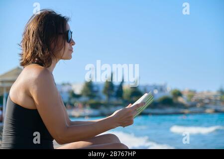Reife Frau in Sonnenbrille Badeanzug Lesebuch auf Sand am Meer sitzend Stockfoto