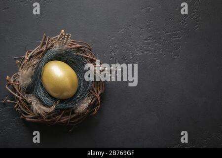 Hähnchen goldenes Ei im Nest auf Schwarz. Oster-Dunkel-Hintergrund. Draufsicht und Kopierbereich. Stockfoto