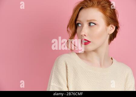 Ein schönes rothaariges Mädchen mit roten Lippen in einem weißen Pullover steht auf einem rosafarbenen Hintergrund und blickt, indem es ihren Kopf dreht, zur Seite Stockfoto