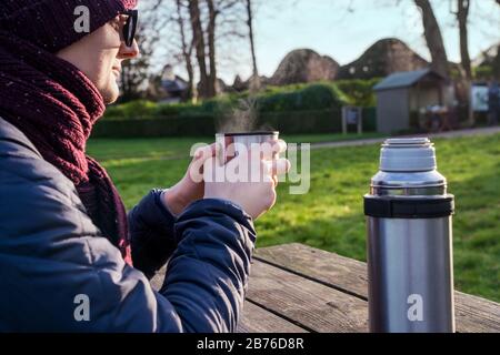 Nahaufnahme kaukasischer junger Reisender Mann in der Sonnenbrille trinkt einen heißen Tee aus Thermos-Tasse mit sonnigen grünen Grasflächen im Freien. Vom Becher mit Tee Stockfoto