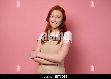 Rothaariges Mädchen in beigefarbener Schürze und T-Shirt steht auf einem rosafarbenen Hintergrund und lächelt. Stockfoto