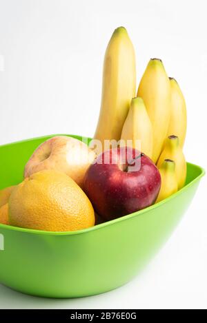 Eine leuchtend grüne Plastikschale mit kunstvoll angeordneten frischen und reifen Früchten wie Äpfeln, Bananen, Orangen und anderen auf einem einfarbigen weißen Hintergrund. Stockfoto