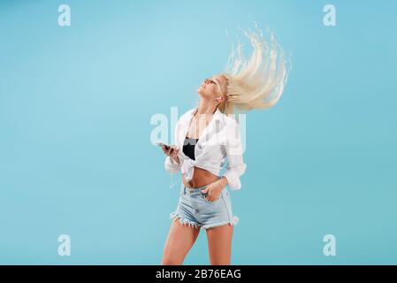 Das junge schlanke blonde Mädchen mit langen Haaren hört Musik auf Kopfhörer und tanzt, wirft den Kopf zurück. Sorgloses Modell im Sommer Kleidung mit Spaß o Stockfoto