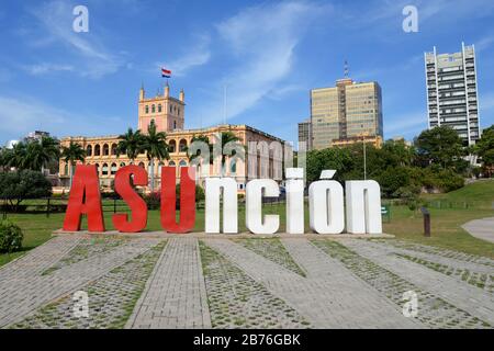 Asuncion Schild vor dem Regierungspalast im Kolonialstil. Park mit Bäumen und Gebäuden in der Innenstadt sichtbar. Beliebter Fotopunkt in Paraguay. Stockfoto