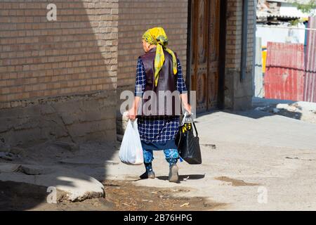 Die alte asiatin, die von hinten gesehen wurde, hält Taschen Handtasche und trägt traditionelle Kleidung mit Stempel und farbenfrohem Schal in Samarkand, Usbekistan. Stockfoto