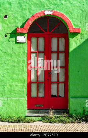 Alte rote Tür mit verrosteter und rissiger Farbe in einer grünen Wand mit Gras im Vorderteil Stockfoto