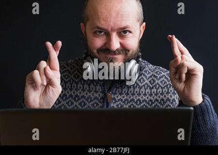 Kaukasischer Erwachsener in Vintage-Pullover und Kopfhörer rund um den Hals hinter dem Laptop, gekreuzte Finger, Kopf und Schultern, schwarzer Hintergrund Stockfoto