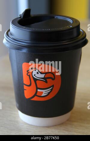 08-02-2020, Russland, Syktyvkar. Kaffeetasse aus schwarzem Papier mit schwarzem Kunststoffdeckel und orangefarbenem Logo der internationalen Pizzeria Kette. Dodo Pizza steht auf dem Tisch in der Nähe des Fensters im Restaurant. Stockfoto