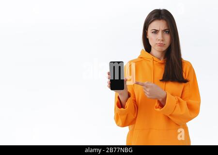 Skeptische und zweifelhafte hübsche kaukasische Mädchen in orangefarbenem Hoodie haben Zweifel, misstrauisch und zögernd zeigende Handyanzeige zu erfroren, gefunden Stockfoto