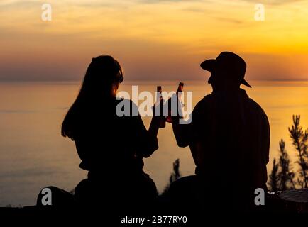 Silhouette eines romantischen Paares, das Bier trinkt und bei Sonnenuntergang auf einer Klippe am Meer sitzt. Stockfoto