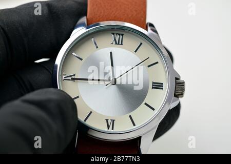 Silberne Armbanduhr mit einem braunen einteiligen Lederriemen. Hand in einem schwarzen glowe hält die Uhr. Weißer Hintergrund. Stockfoto