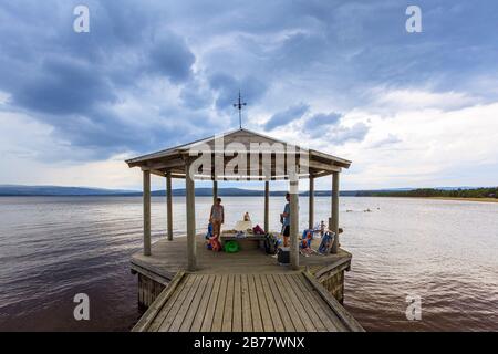 Eine Familie, die in einem Pavillon am Ende eines kleinen Piers Schutz sucht, während sich am Horizont Gewitterwolken sammeln. Stockfoto