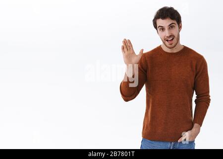 Hallo. Gutaussehender kaukasier 25er Kerl in Sweatshirt, Hand in der Tasche halten und locker informell grüßen Person, lächeln und winken erhabene Hand in Hallo Stockfoto