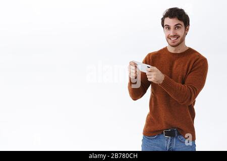 Fröhlicher lächelnder junger 25-er Mann mit Bart mit Smartphone zum aufnehmen von Videos, zum anschauen von Live-Streams online, zum horizontalen halten des Mobiltelefons und zum Spielen Stockfoto