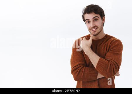 Taille-up-Porträt von interessiertem gut jungen Mann hören neugierige Nachrichten, lächeln und sehen fasziniert, berühren Kinn nachdenklich, erwägen Kauf Stockfoto