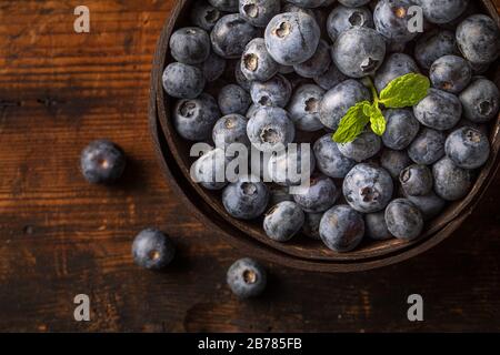 Blaubeeren oder Hucklebeeren und Minzblatt in einer Kokosschale, von oben gesehen. Die Schüssel befindet sich auf einem rustikalen Holztisch. Es gibt einige Blaubeeren außerhalb Stockfoto