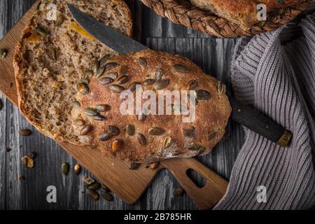 Hausgemachtes Brot mit Aprikosen und Kürbissamen. Der Bun wird mit einem Vintage-Messer zur Hälfte geschnitten und befindet sich auf einem Holzschneidbrett. Von oben gesehen, Stockfoto