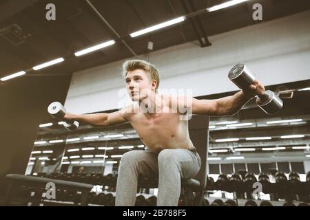 Konzentrierter sportlicher Kerl beim Workout im Fitnessstudio mit Hanteln. Hebt schwere Gewichte, trainiert die Bizeps. Schöne Brustmuskeln. Stockfoto