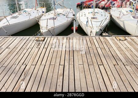 Vier kleine Lernsegelboote hintereinander in einem Holzsteg. Großer Kopierraum zum Platzieren von Text und einigen roten pneumatischen Beibooten im Hintergrund. Stockfoto