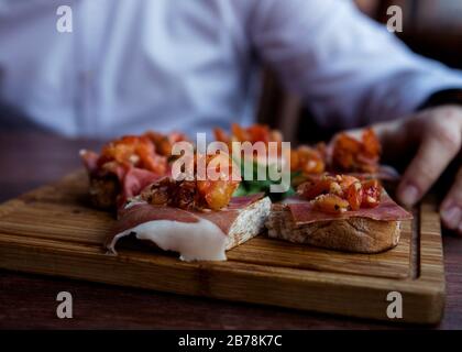 Spanisches Tapas wird im Restaurant auf rustikalem Holzbrett serviert. Bruschetta mit Prosciutto, Tomaten, Ruccola und Gewürzen Stockfoto