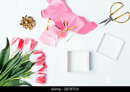 Ungepackte leere Geschenkbox neben zerrissenem Verpackungspapier, Band, Schleife und Blumenstrauß von Tulpen auf weißem Tisch, Blick von oben. Stockfoto