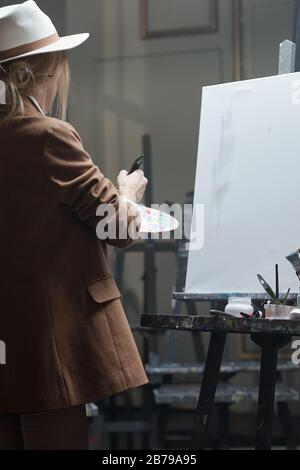 Junge Frau in Hut und Jacke, die Farben auf Palette mischt, während sie mit leerem Papier vor der Staffelei steht und malen will Stockfoto