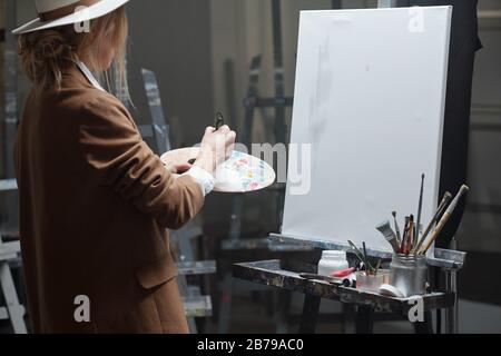 Junge, zeitgenössische Frau mit Farbpalette, die Farben mischt, während sie im Studio mit leerem Papier auf Staffelei malen will Stockfoto