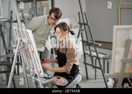 Ziemlich fleißiges Mädchen in der Schürze, das bei der Lektion über das Malen arbeitet und dem Rat ihres Lehrers folgt, der in der Nähe steht Stockfoto