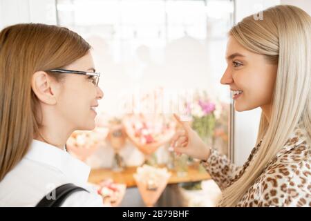 Hübsche junge blonde Frau, die ihren Freund ansieht, während sie beide vor der Anzeige des Blumenhandels stehen und über Blumen diskutieren Stockfoto