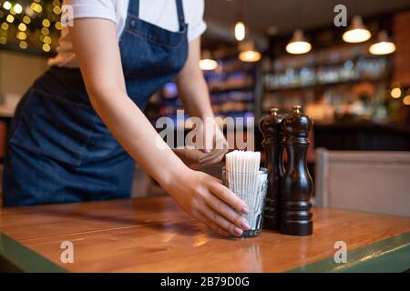 Junge Kellnerin in der Schürze, die Glas mit Zahnstocher auf Holztisch legt, während sie sich für neue Gäste im Café oder Restaurant vorbereitet Stockfoto