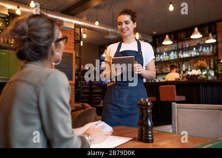 Fröhliche junge Kellnerin mit digitalem Tablet, das an einem Tisch vor weiblichem Gast steht und ihre Bestellung im Café oder Restaurant abnimmt Stockfoto