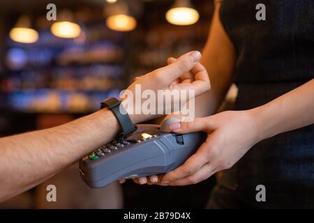 Hand des jungen Kunden von Café oder Restaurant, der mit Smartwatch über den Zahlungsautomaten bewaffnen bleibt, während er für Getränke oder Essen bezahlt Stockfoto