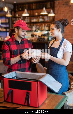 Der junge Kurier in der Arbeitskleidung, der Plastikbehälter mit Lebensmitteln aus den Händen der Kellnerin mitnimmt und ihm hilft, Bestellungen von Kunden im Café zu verpacken Stockfoto