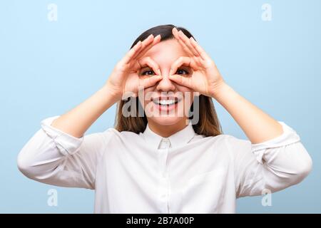 Sehhilfen: Junge Frau hält ihre Hände über ihre Augen und imitiert Brille und lächelt fröhlich vor blauem Hintergrund Stockfoto
