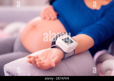 Schwangere Frauen überwachen ihren Blutdruck zu Hause mit dem medizinischen Gerät am Handgelenk und legen ihren Bauch frei. Stockfoto