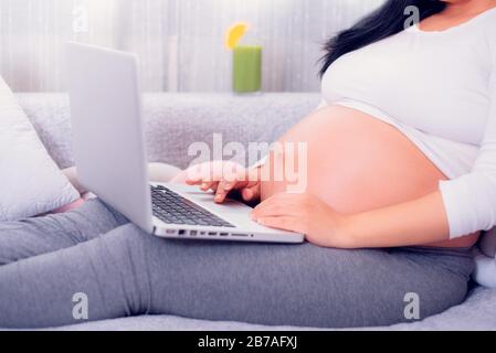 Schwangere Frau, die ihren Laptop benutzt, während sie sich zu Hause auf der Couch entspannt und ihren schwangeren Bauch aussetzt. Stockfoto