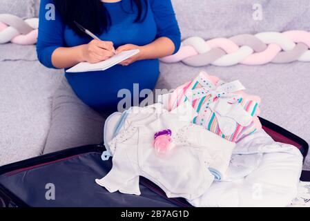 Schwangere Frau, die die Checkliste durchgeht, während sie den Koffer mit neugeborenem Babykleidung für das Krankenhaus packt. Stockfoto