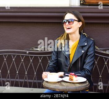 Mädchen in Sonnenbrille in der Nähe von leckeren Beerenkuchen und Kaffee auf dem Tisch auf brauner Terrasse Hintergrund. Die Dame trinkt während der Kaffeepause Kaffee. Konzept für Patisserie und Pausenzeit. Frau mit glücklichen Gesichten sitzt im Café Stockfoto