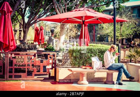 Frau, die an einem sonnigen Tag auf einer Bank vor der Restaurantterrasse des trendigen Restaurants Olive & Ivy in Scottsdale, AZ, USA mit Handy sitzt Stockfoto