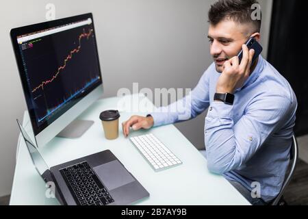 Lächelnder junger männlicher Online-Händler, der mit dem Kunden auf dem Smartphone spricht, während er einen Laptop am Schreibtisch verwendet Stockfoto