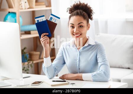 Afrikanische Touragentin mit weiblicher Abfahrt sitzt am Tisch im Büro und hält Pässe und Flugtickets, die die Kamera lächelnd fröhlich aussehen Stockfoto