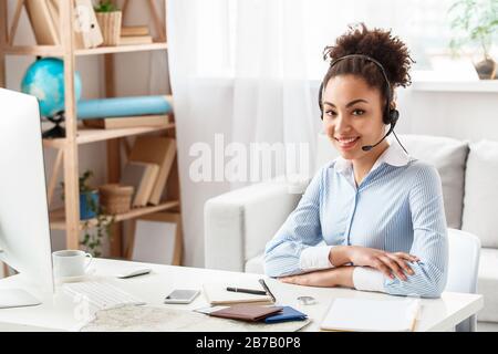 Afrikanische Touragentin mit weiblichem Abstieg, die am Tisch im Büro sitzt und ein Headset trägt, das positiv lächelnd auf die Kamera blickt Stockfoto