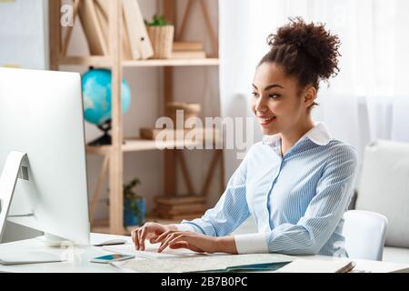 Afrikanische Abstammung, weibliche Touragentin sitzt am Tisch im Büro und tippt Buchungsinformationen auf dem Computer lächelnd fröhlich konzentriert Stockfoto