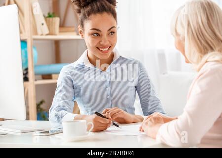 Afrikanischer Abstieg weiblicher Reisebüro im Büro lächelte fröhlich und erklärte Bedingungen für eine ältere Frau, die einen Vertrag unterzeichnet Stockfoto