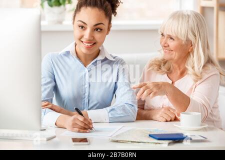 Afrikanische Abfahrerin, weibliche Reisevermittlerin im Büro mit einer älteren Frau, die einen Computermonitor ansieht, Notizen macht, lächelnd glücklich über die Reise Stockfoto