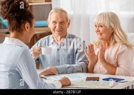Afrikanische Abstiegsfrau Reisebüro im Büro sitzt am Tisch mit einem Paar, das heiße Kaffees trinkt und über die zukünftige Reise lächelnd fröhlich diskutiert Stockfoto