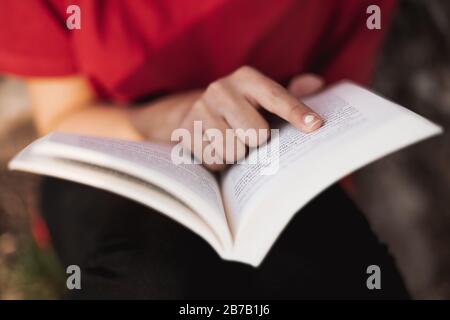 Nahaufnahme eines offenen Buches direkt in der Mitte von einer jungen Frau, die mit dem Finger in ein Wort der Seite zeigt. Geringe Schärfentiefe.