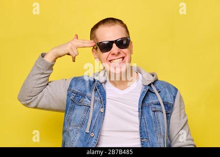 Stilvoller kaukasischer Mann in einer Jeans und Sonnenbrille auf gelbem Hintergrund. Nahaufnahme. Fasst abstrakt eine Hand in der Nähe seines Kopfes wie eine Pistole. Stockfoto